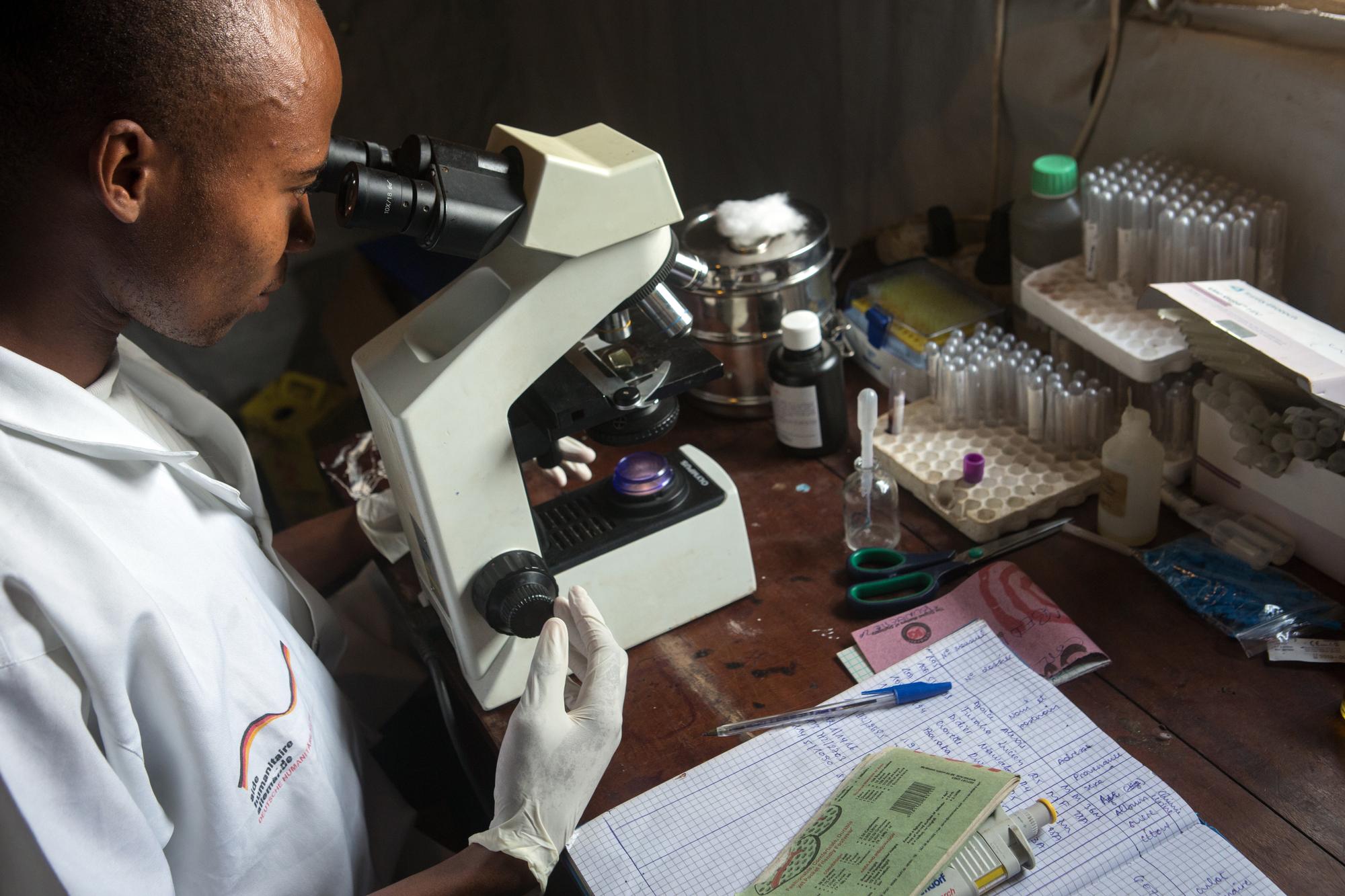 A lab technician at a wooden desk looks into a microscope.